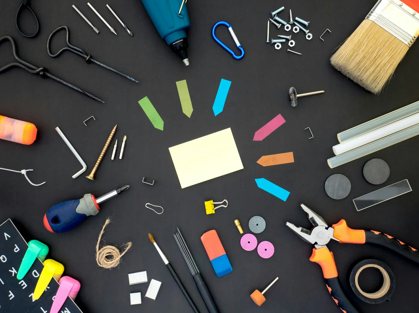 Different tools against a black background, including a screwdriver, drill, paintbrushes, colourful post-its, staples, eraser, and more.